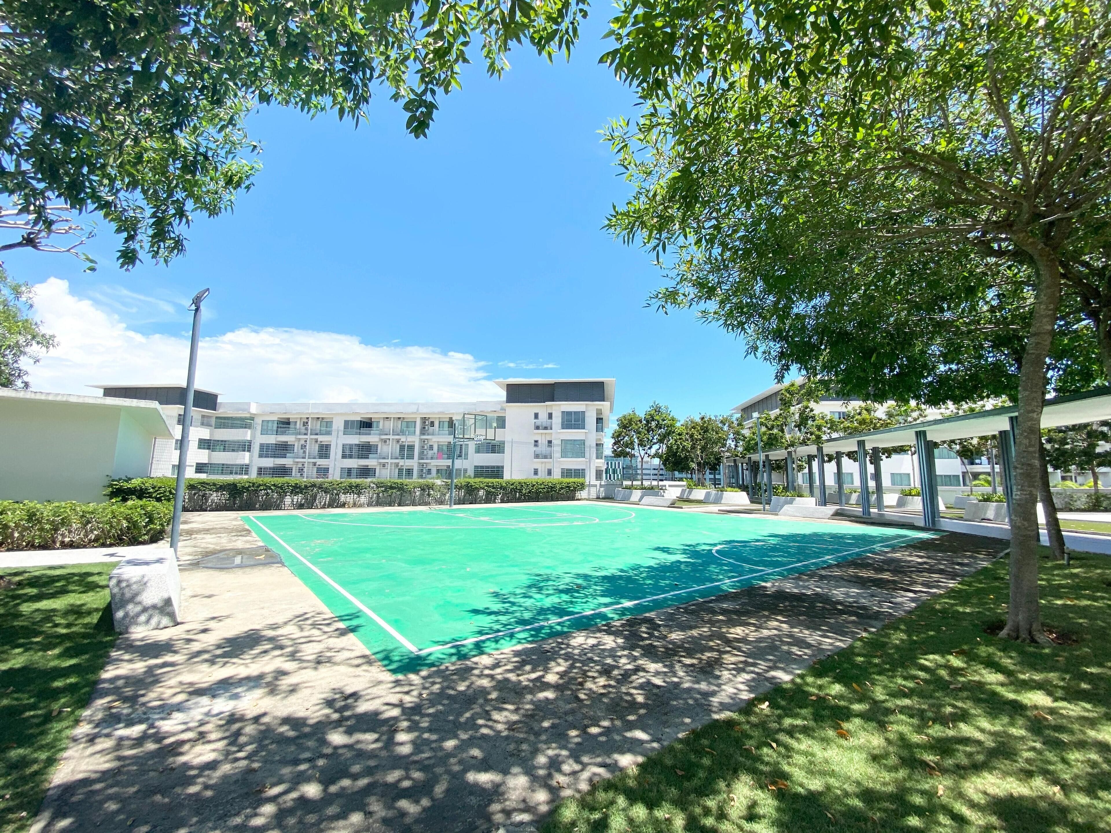 A basketball court with a green surface, surrounded by trees and a building with multiple windows in the background.
