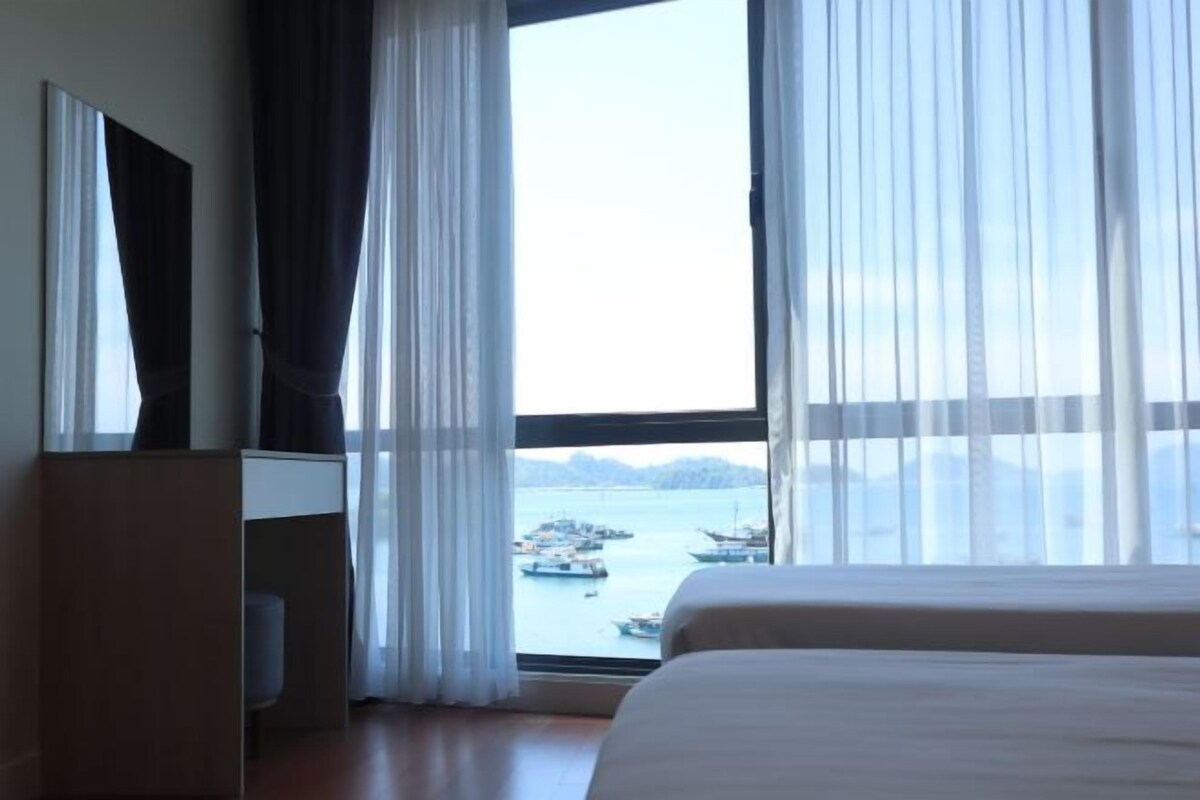 Quarto de hotel com uma cama grande, uma televisão sobre um suporte e vista para barcos na água através de cortinas translúcidas.