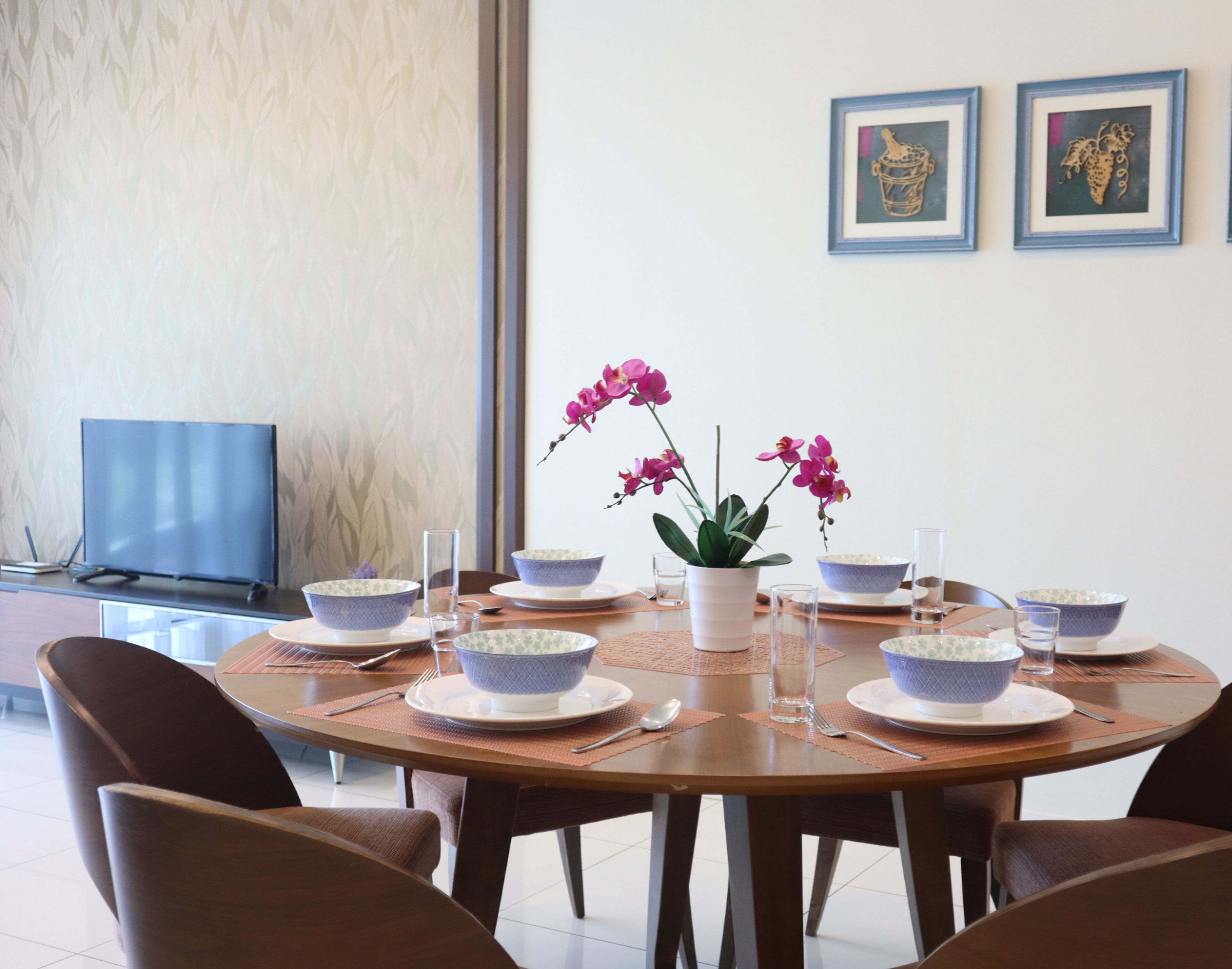 A dining table set with bowls, plates, and cutlery, surrounded by chairs, with a potted plant and framed artwork on the wall.