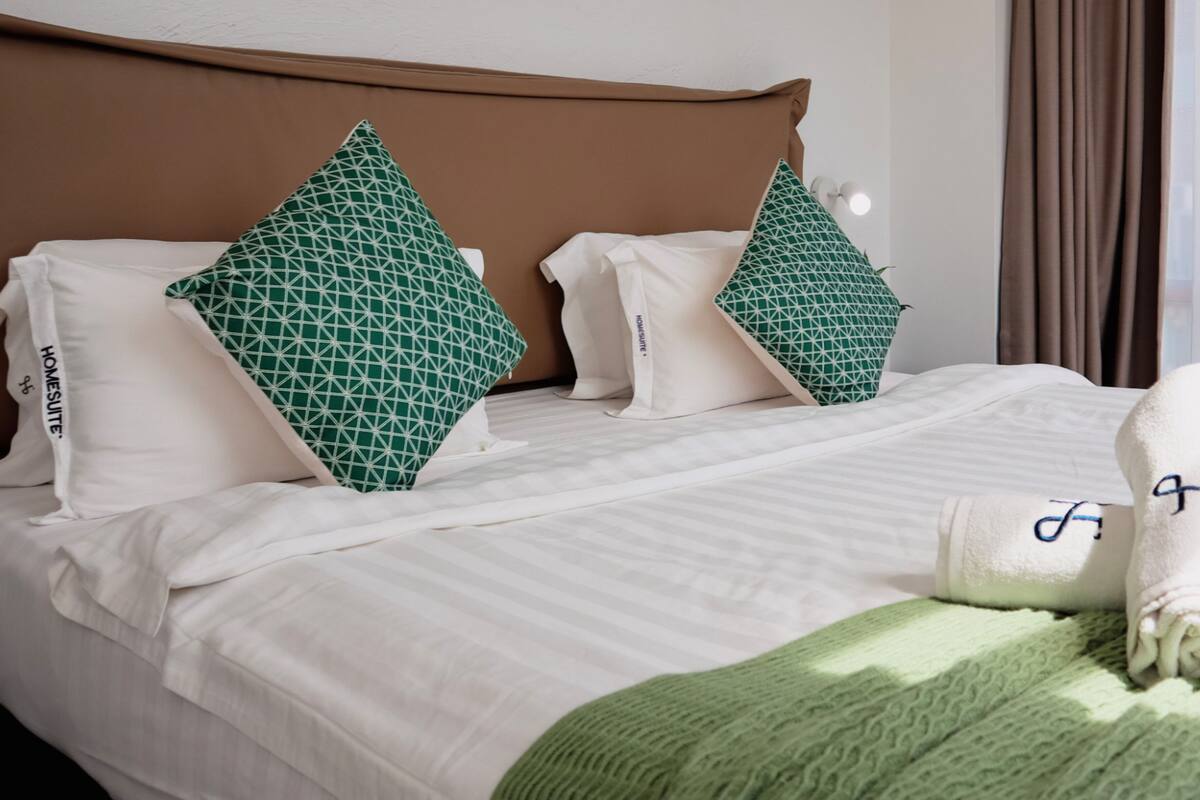 A neatly made bed with white linens and two green patterned pillows, a brown headboard, and a green throw blanket.