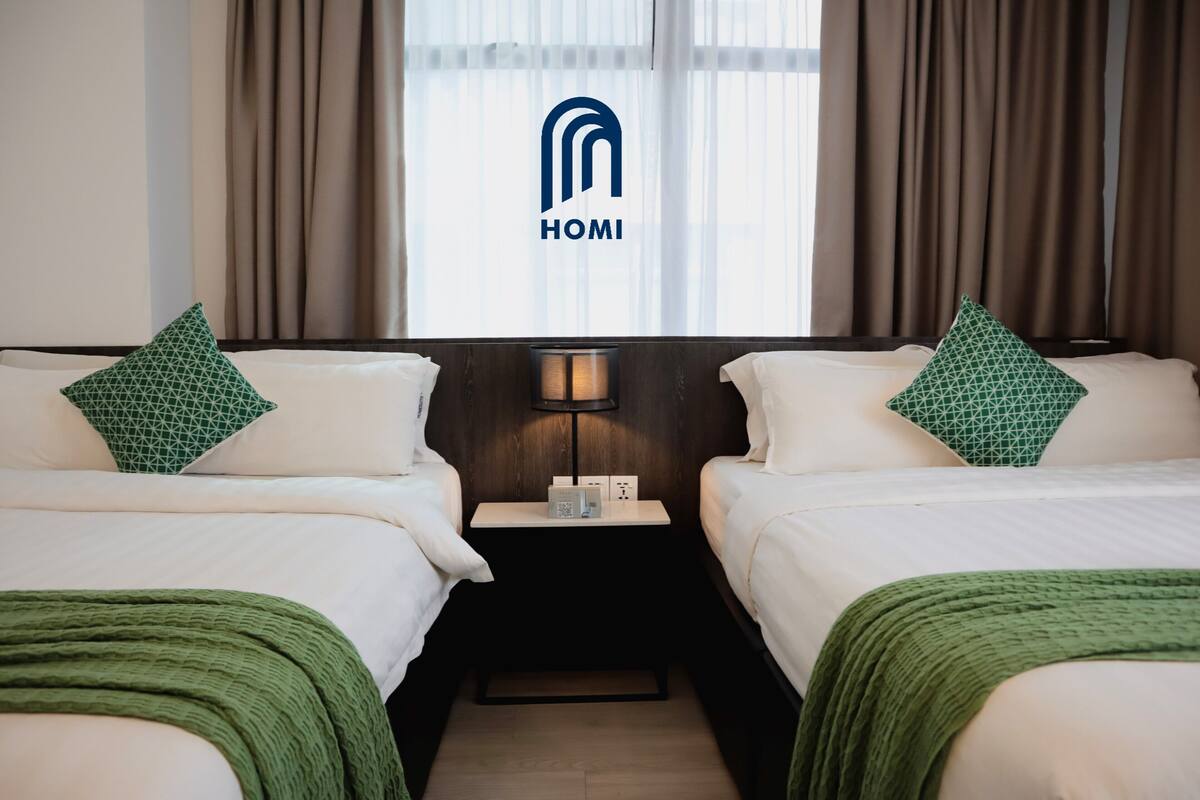 A hotel room with two beds, each with a green patterned pillow, a bedside table with a lamp, and a window with curtains.