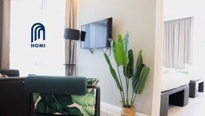 A modern hotel room with a flat-screen TV mounted on the wall, a large potted plant, a bed with white bedding, and a dark-colored armchair with a green cushion. - Homi Oasis 和逸绿  洲 (Kota Kinabalu)