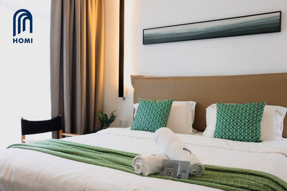 A hotel room with a bed, green and white patterned pillows, a green throw blanket, and a folded towel.