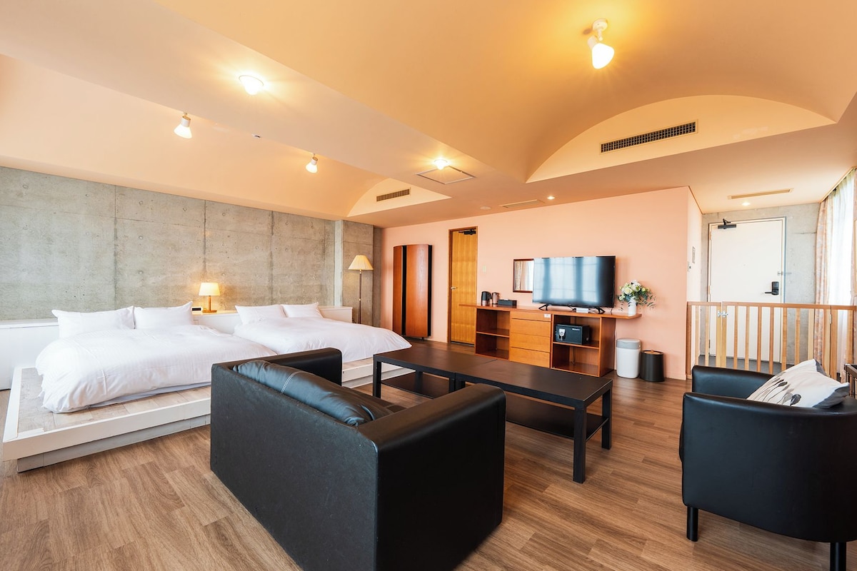 A modern hotel room with a large bed, a desk with a TV, and a seating area with chairs and a sofa.