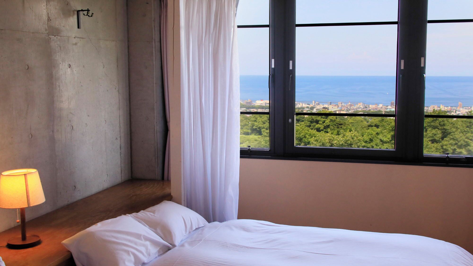 Une chambre à coucher avec un lit, une lampe, une fenêtre donnant sur la ville et la mer, et un rideau.