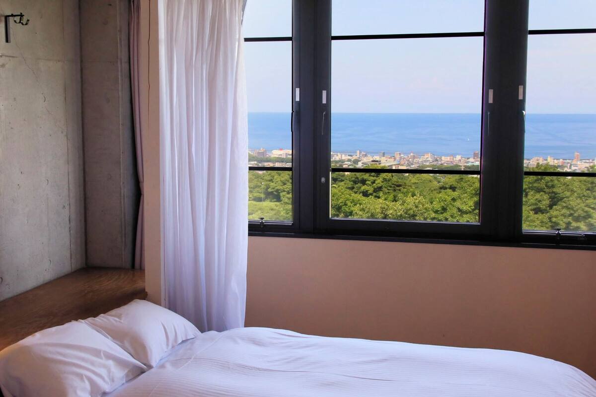 Quarto com cama, luminária, janela com vista para a cidade e mar, e cortina.