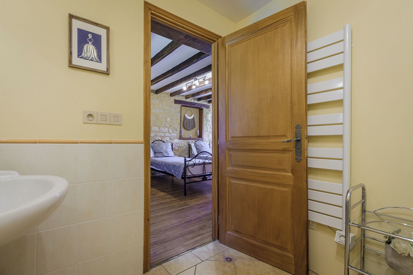 Un baño con un lavamanos blanco, una puerta de madera que da a un dormitorio con cama y pared de piedra.