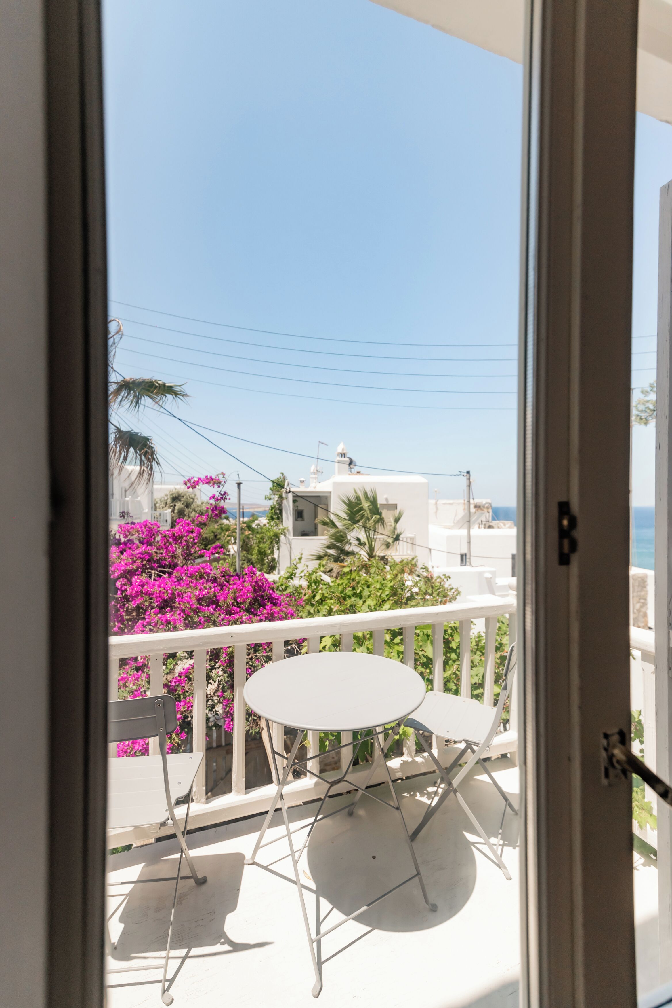 A balcony with a white table and chairs, overlooking a residential area with white buildings and pink bougainvillea flowers.