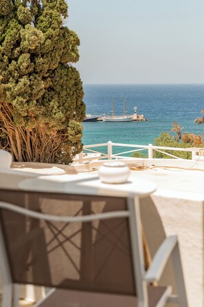 A terrace with a table and chairs, overlooking a calm sea with boats and a lighthouse. - Rania Apartments Sea View (Mykonos)