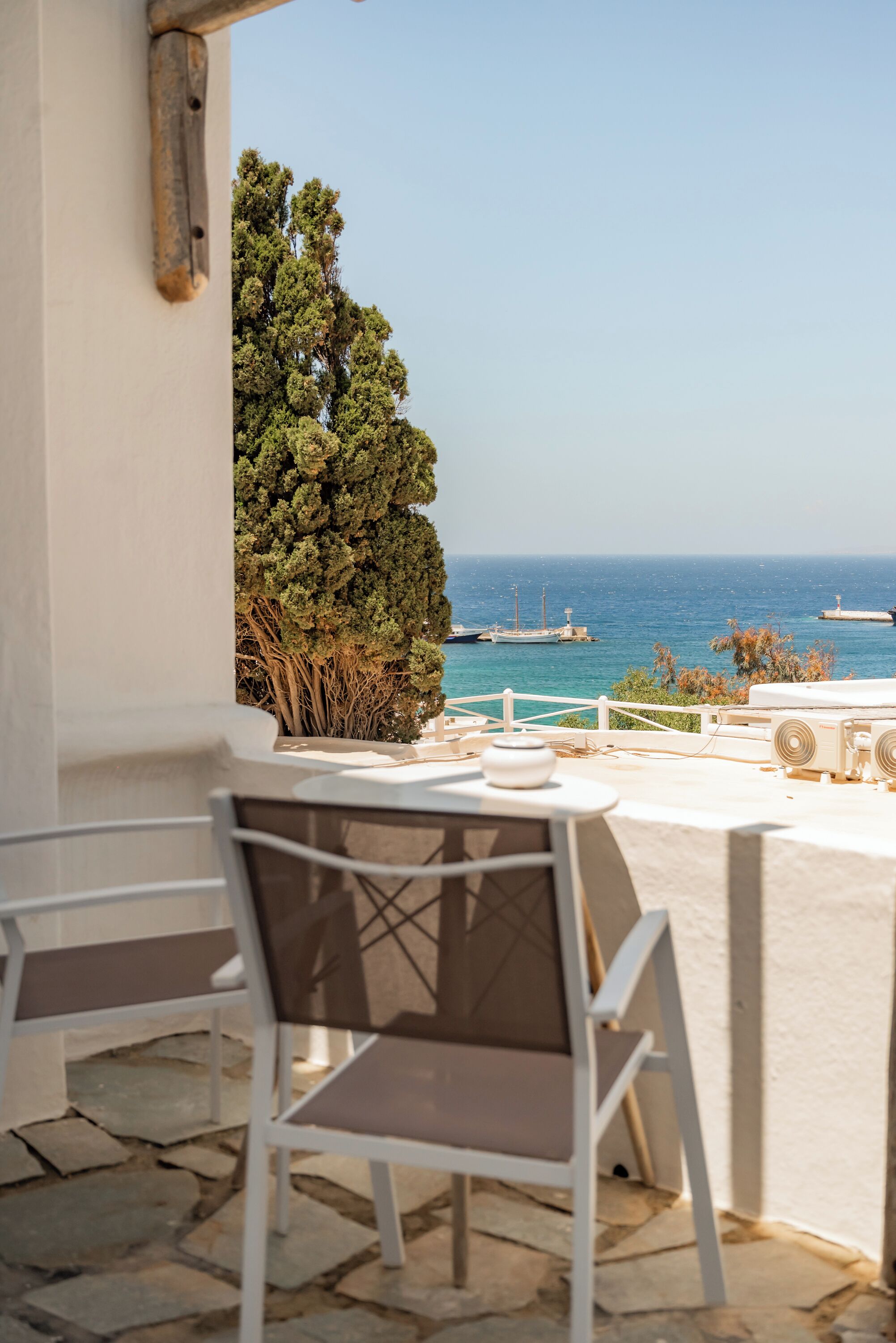Una terraza con una mesa y sillas, con vistas a un mar azul y despejado y un árbol.