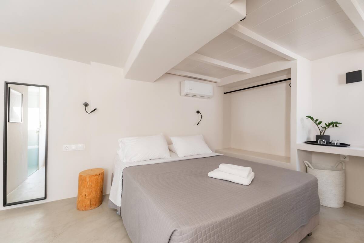 A modern bedroom with a large bed, a wooden stool, a mirror, and a wall-mounted air conditioner.