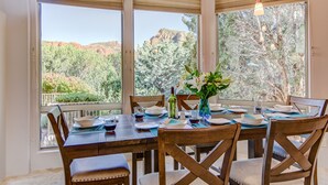 A dining area with a table set for four, large windows with blinds, and a view of a mountainous landscape. - Sedona Spectacular: Luxury 3BR home with private pool, hot tub, and stunning red rock views. (Sedona)