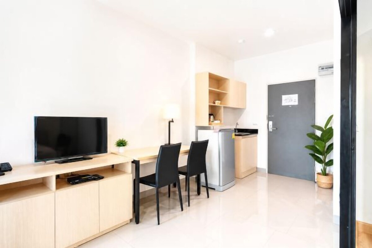 A modern, well-lit room with a flat-screen TV, a desk, two chairs, a small plant, and a refrigerator.