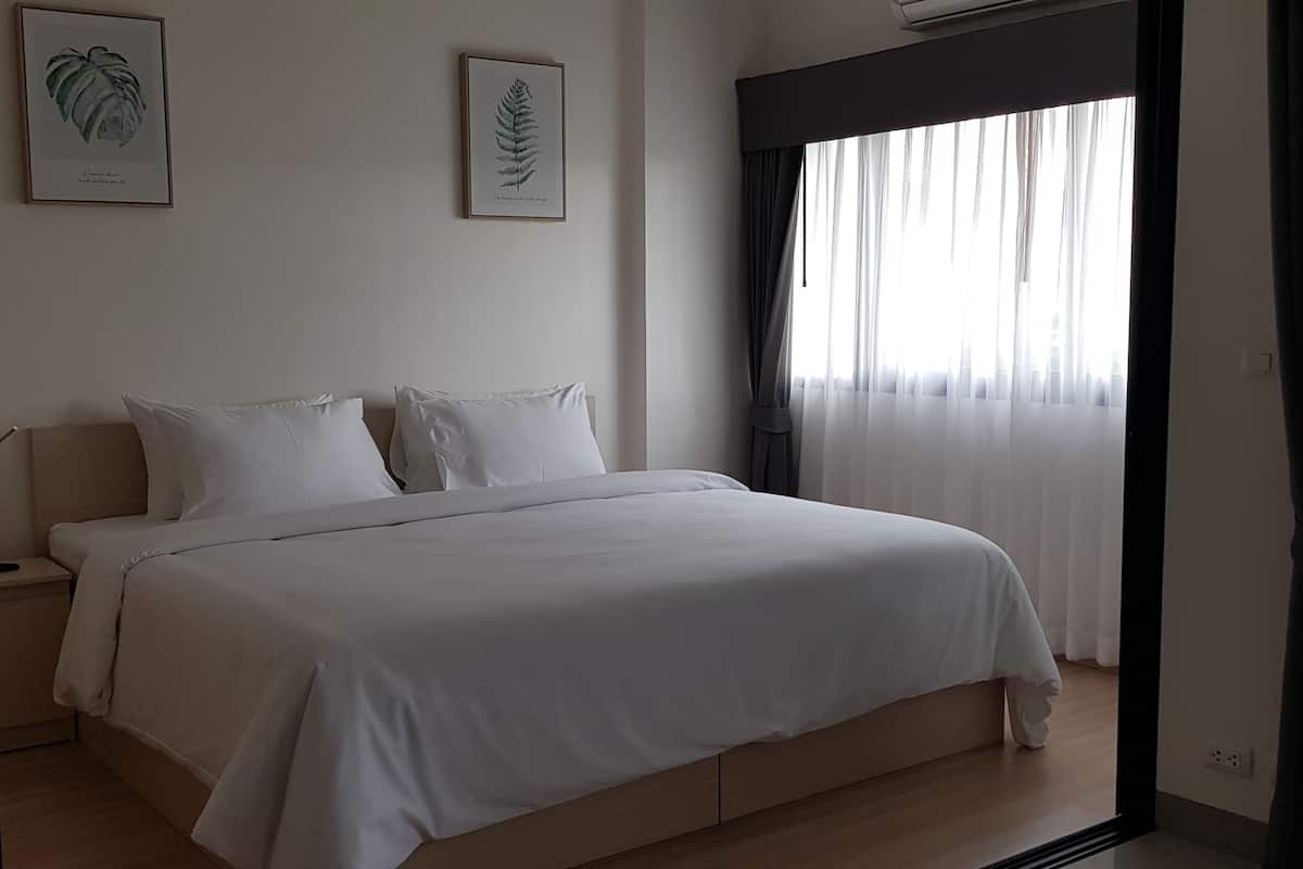 Un dormitorio con cama, dos almohadas, una mesita de noche, lámpara y ventana con cortinas.