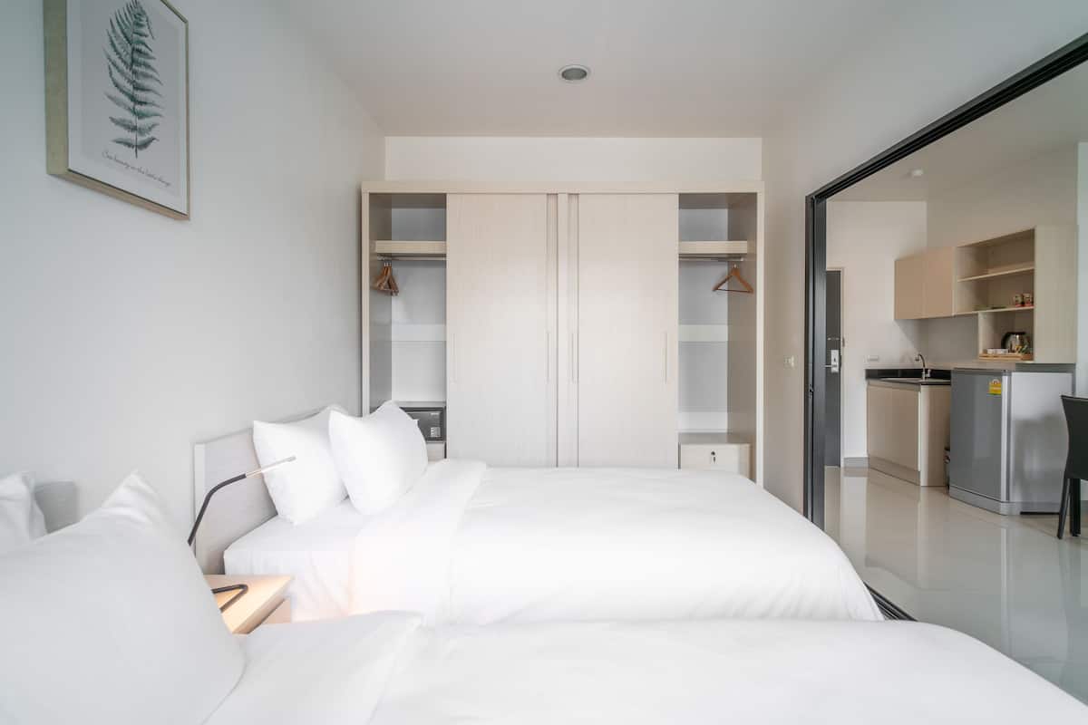 Una habitación de hotel moderna con una cama grande, una mesita de noche, un armario y una cocina visible a través de una puerta abierta.