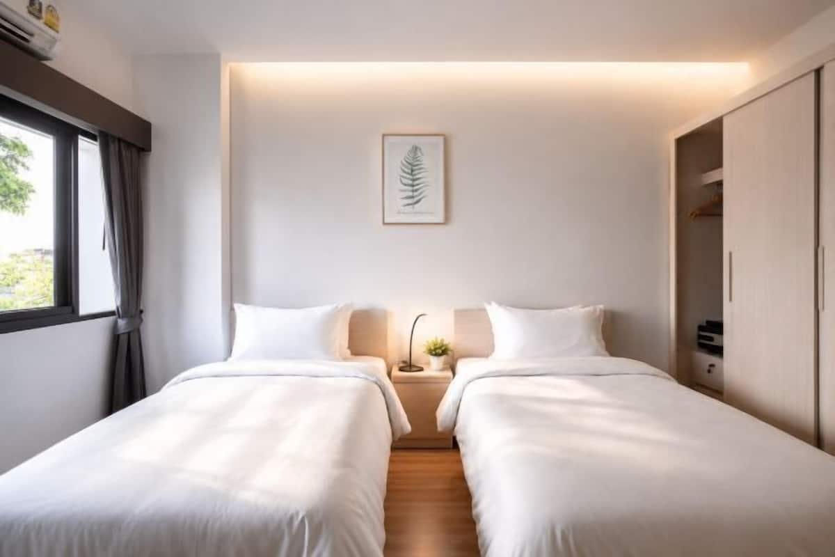 Habitación doble con dos camas, una mesita de noche con lámpara, un cuadro con hojas en la pared y una ventana con cortinas.