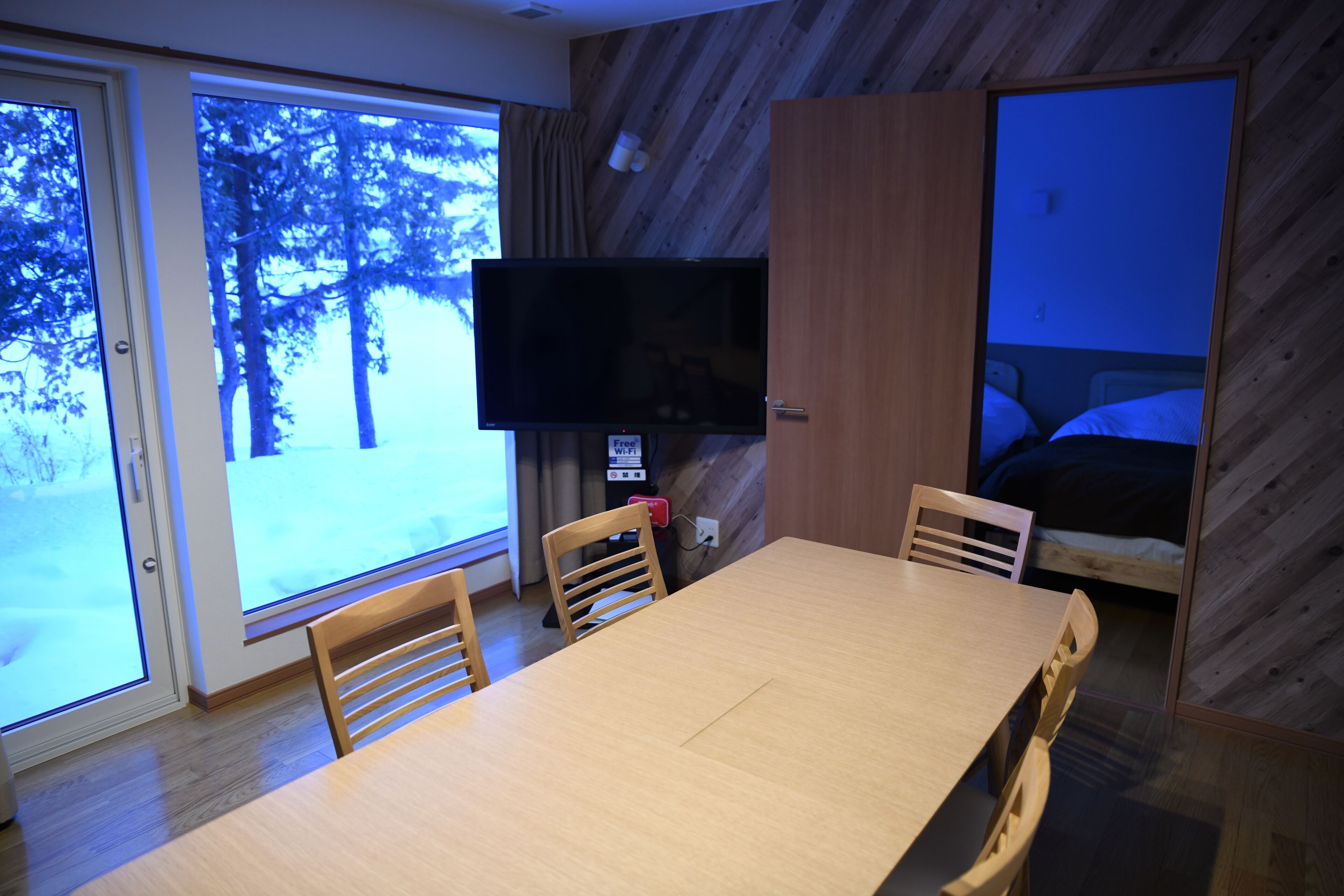 A room with a wooden table, chairs, a flat-screen TV, and a bed.
