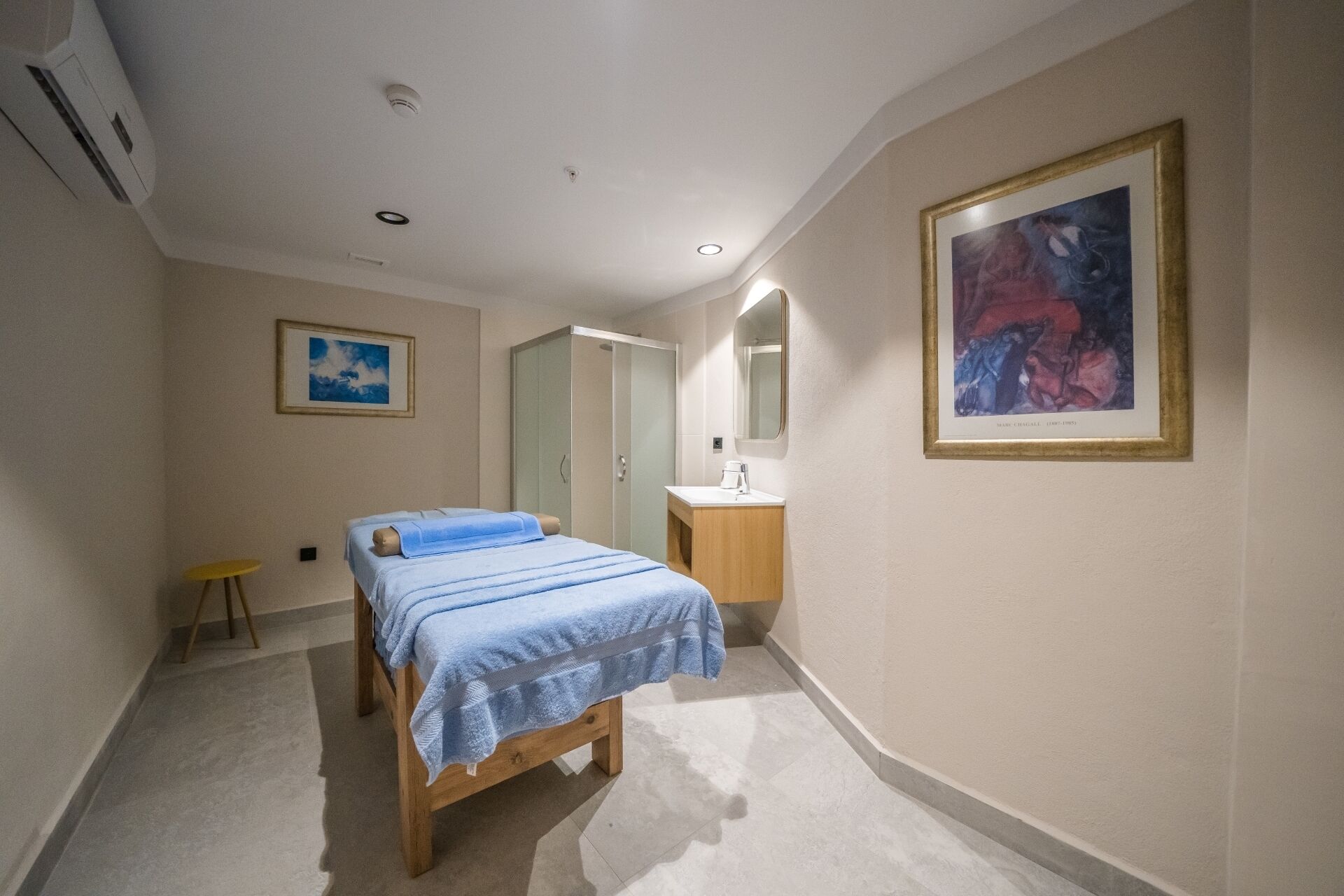 Un dormitorio con cama, mesita de noche, taburete pequeño, baño con lavamanos y espejo, y un cuadro enmarcado en la pared.