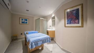 Un dormitorio con cama, mesita de noche, taburete pequeño, baño con lavamanos y espejo, y un cuadro enmarcado en la pared.