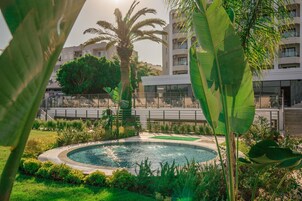 A circular pool surrounded by lush greenery and palm trees, with a modern building in the background.