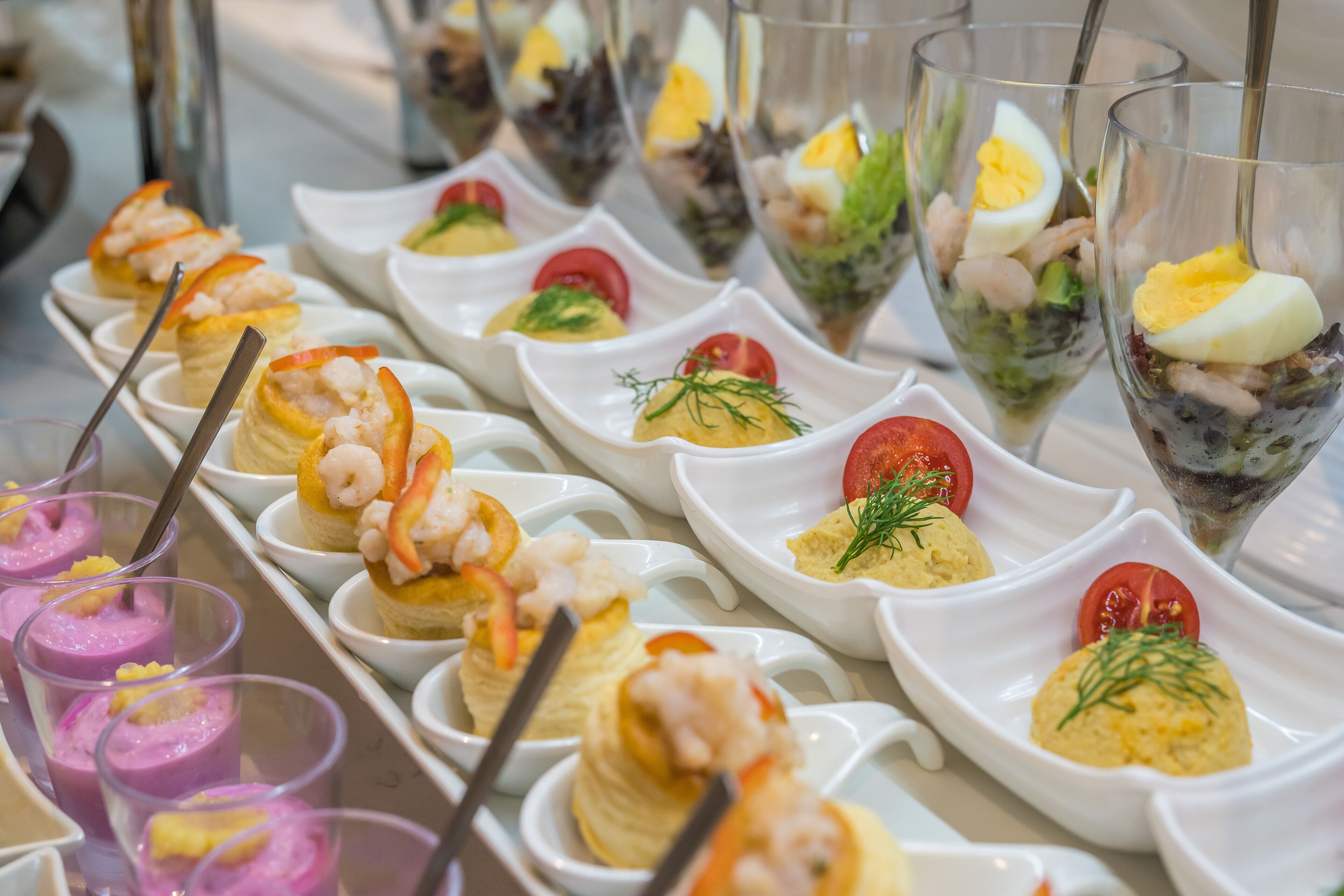Diversos aperitivos, como camarones, huevos y verduras, dispuestos en platos y vasos blancos.