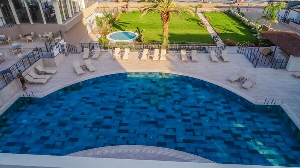 Una gran piscina exterior con un diseño geométrico de azulejos, rodeada de tumbonas y un jacuzzi circular pequeño.