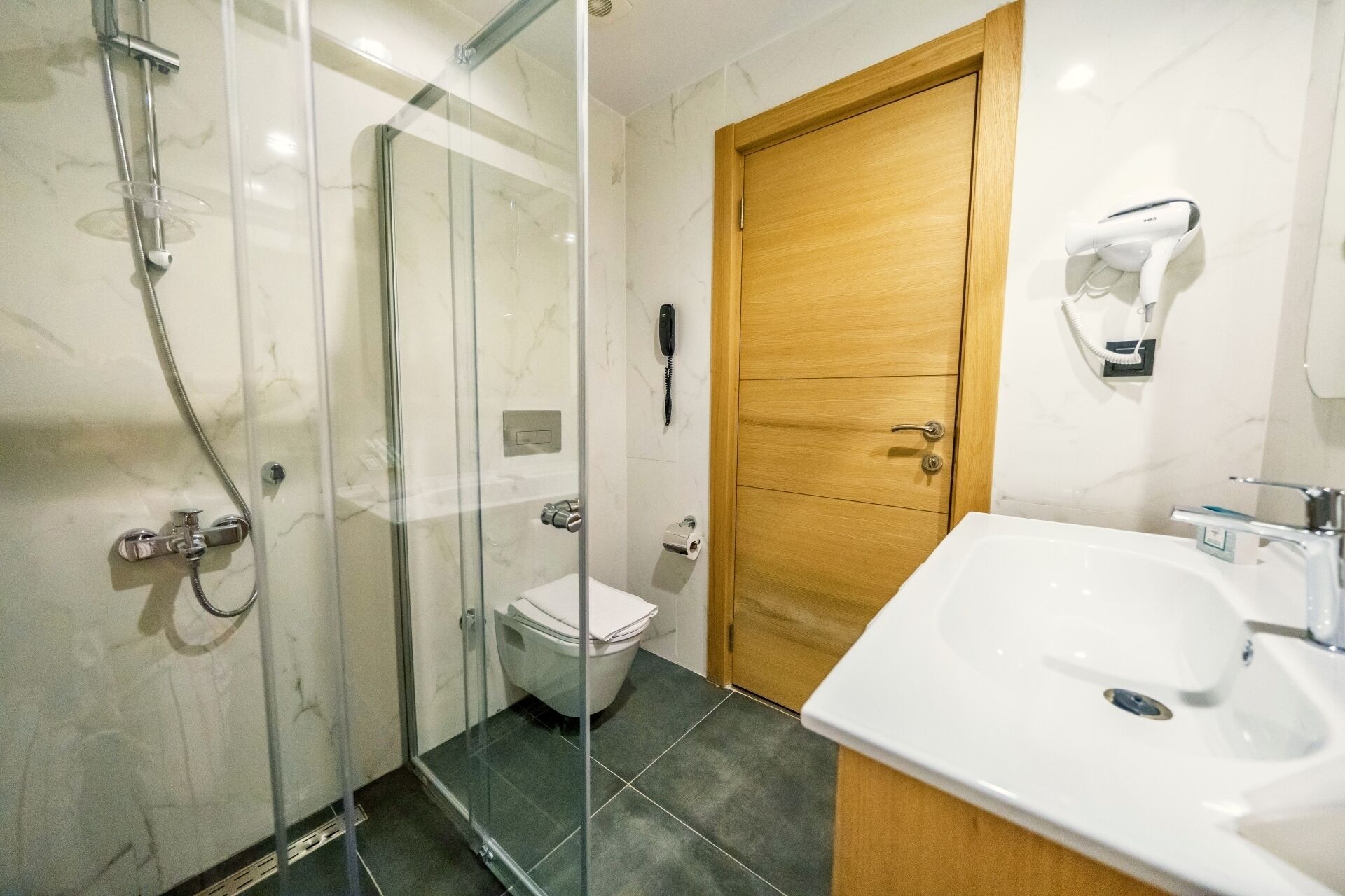 A bathroom with a glass-enclosed shower, a toilet, a wooden door, and a white sink.