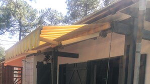Une terrasse extérieure avec une table et des chaises, un espace abrité avec un auvent jaune et une grande porte noire.