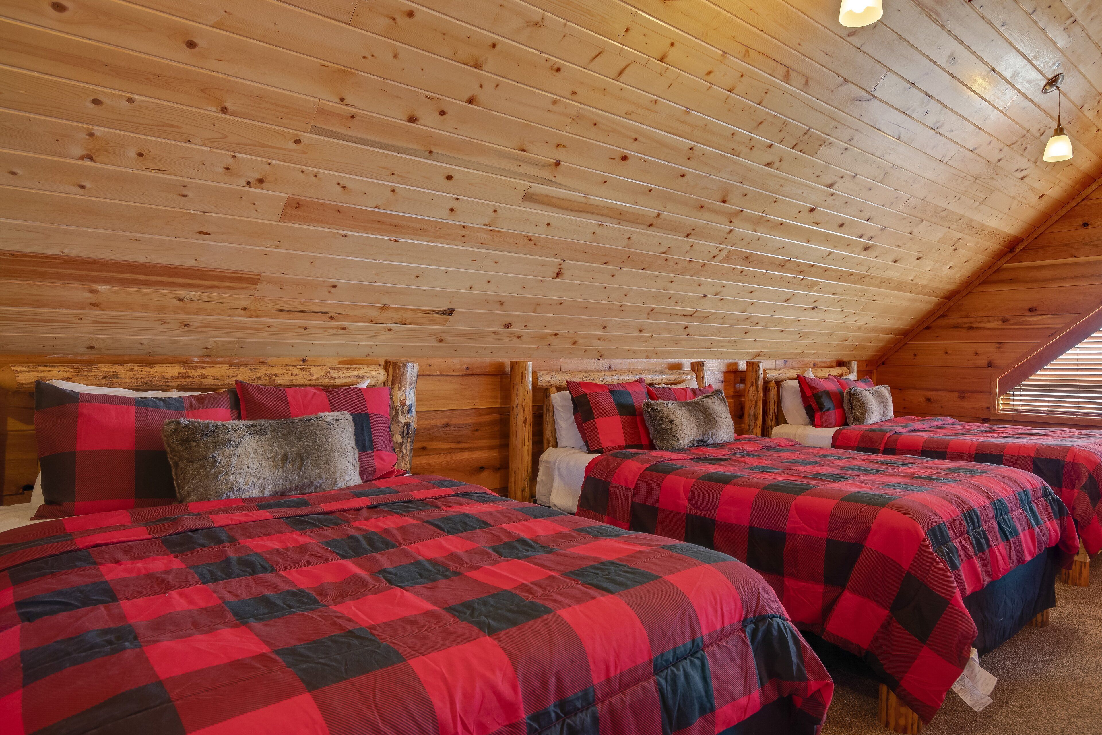 Una cabaña con tres camas, cada una con ropa de cama a cuadros rojos y negros, paredes de madera y techo inclinado con luces colgantes.