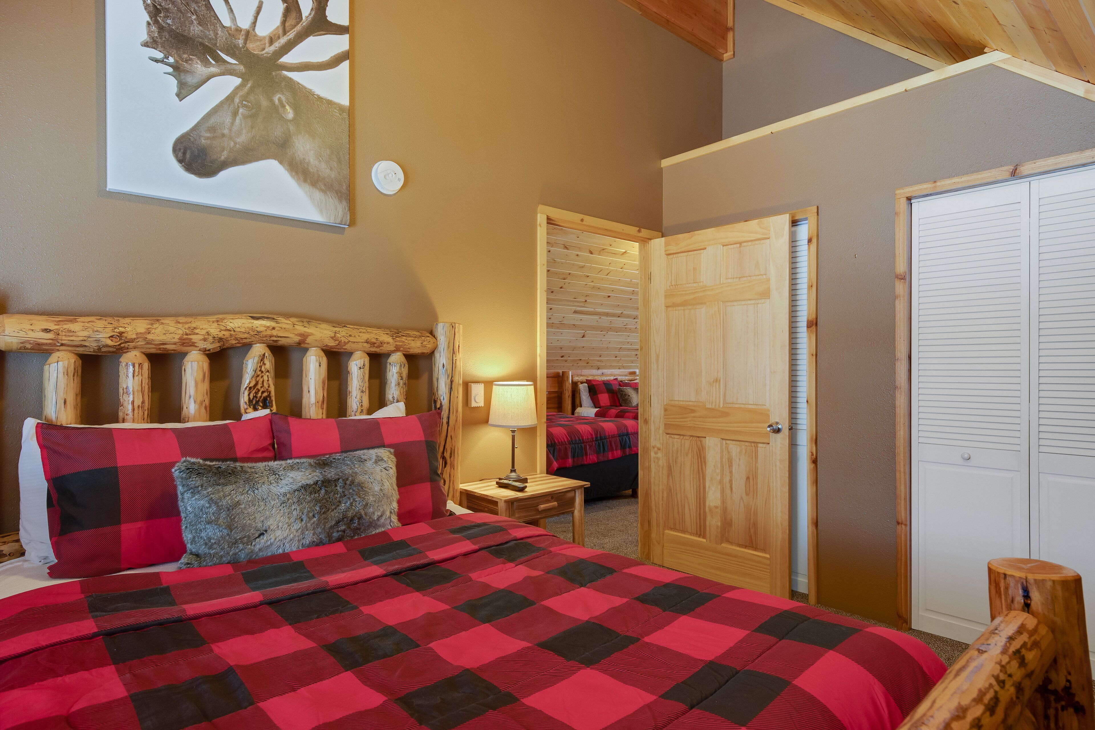 A bedroom with a large bed, a wooden headboard, a red and black checkered bedspread, and a deer head painting on the wall.