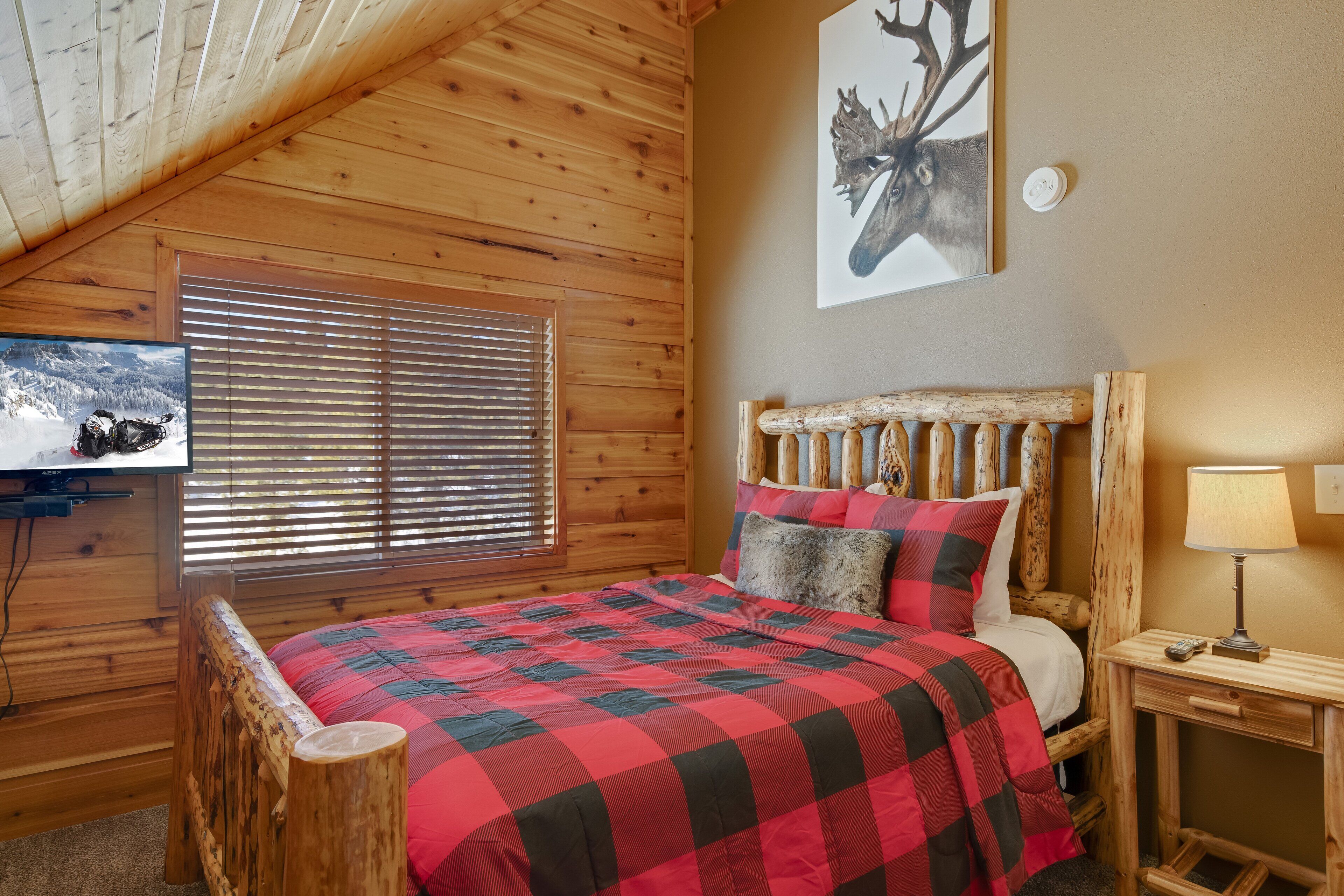 A wooden cabin bedroom with a bed, a wooden headboard, a nightstand, a lamp, and a television on the wall.