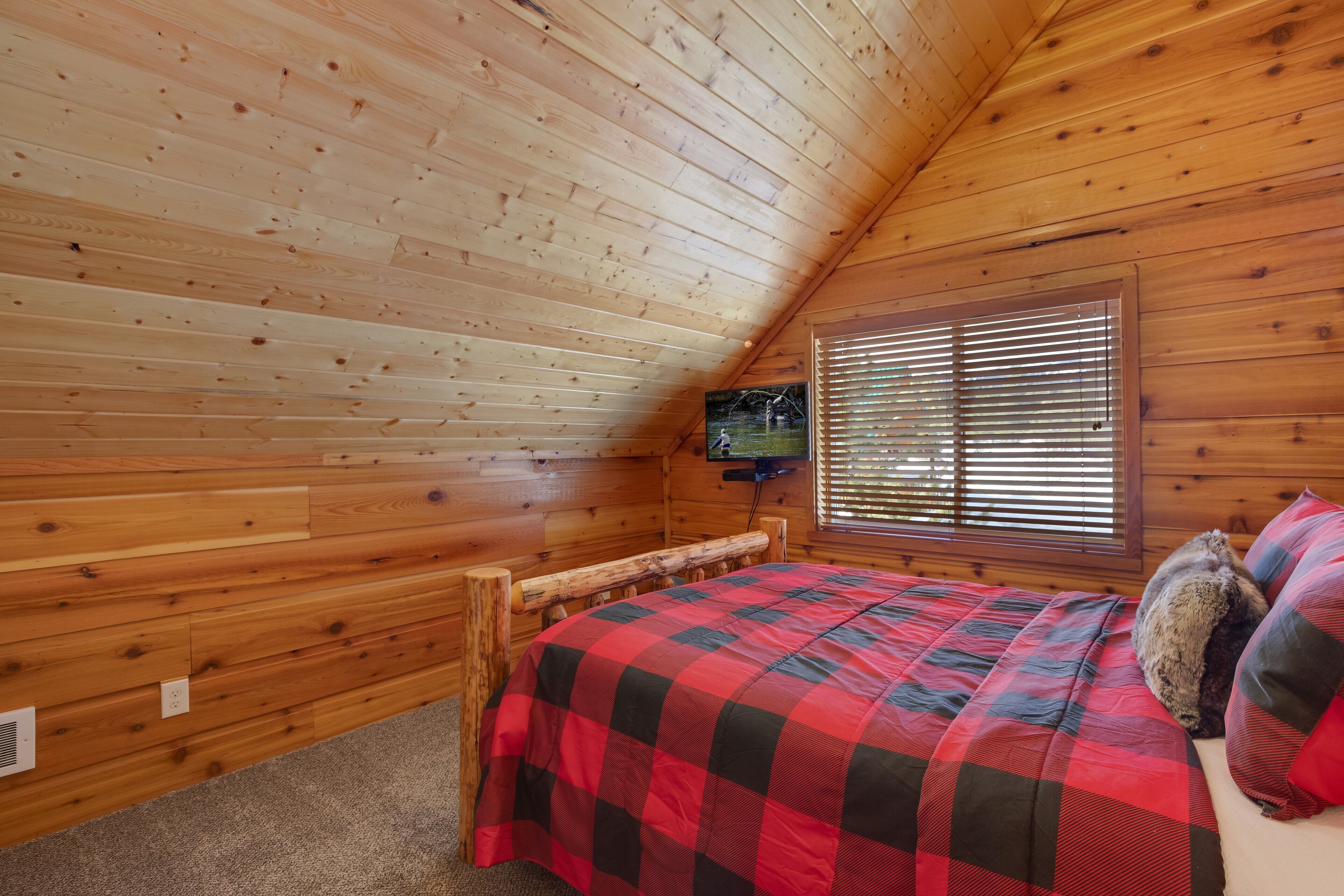 Een gezellig houten kamer met een bed, een raam met zonwering, en een aan de muur bevestigde televisie.
