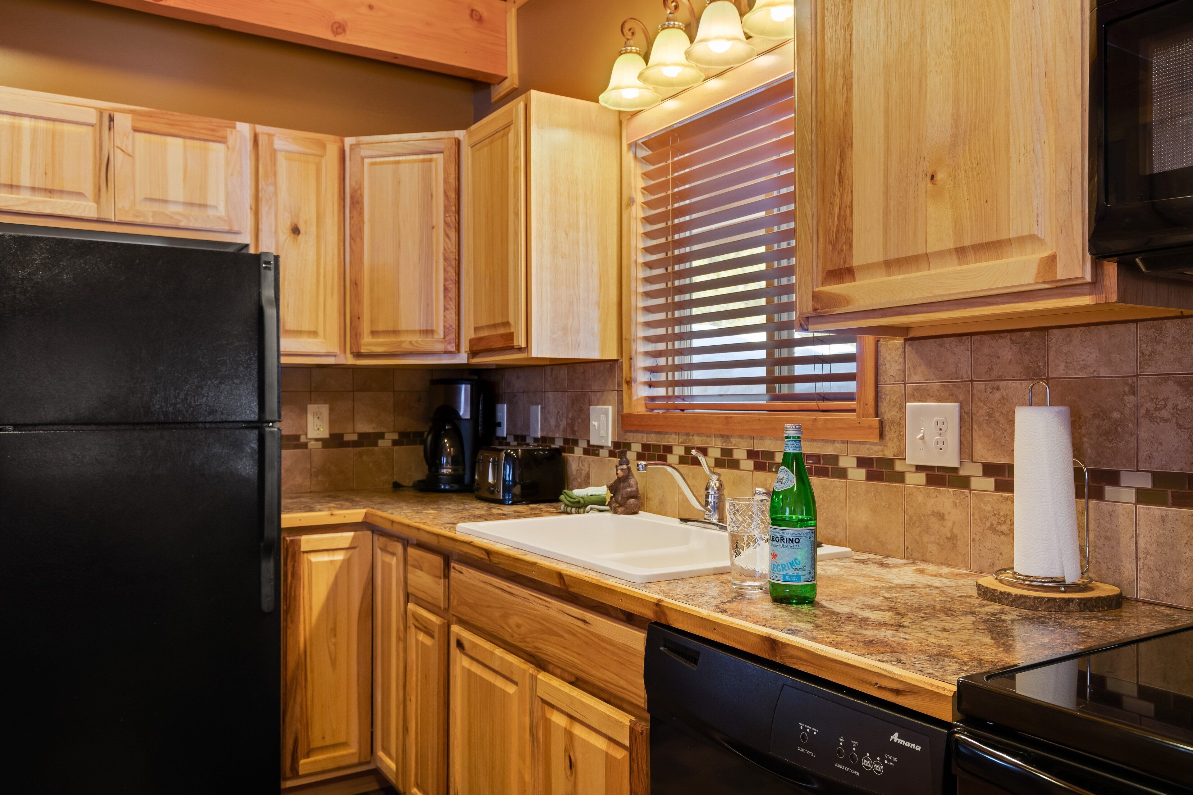 Una cocina con armarios de madera, un refrigerador negro, un fregadero y una encimera con una botella de cerveza y un vaso.
