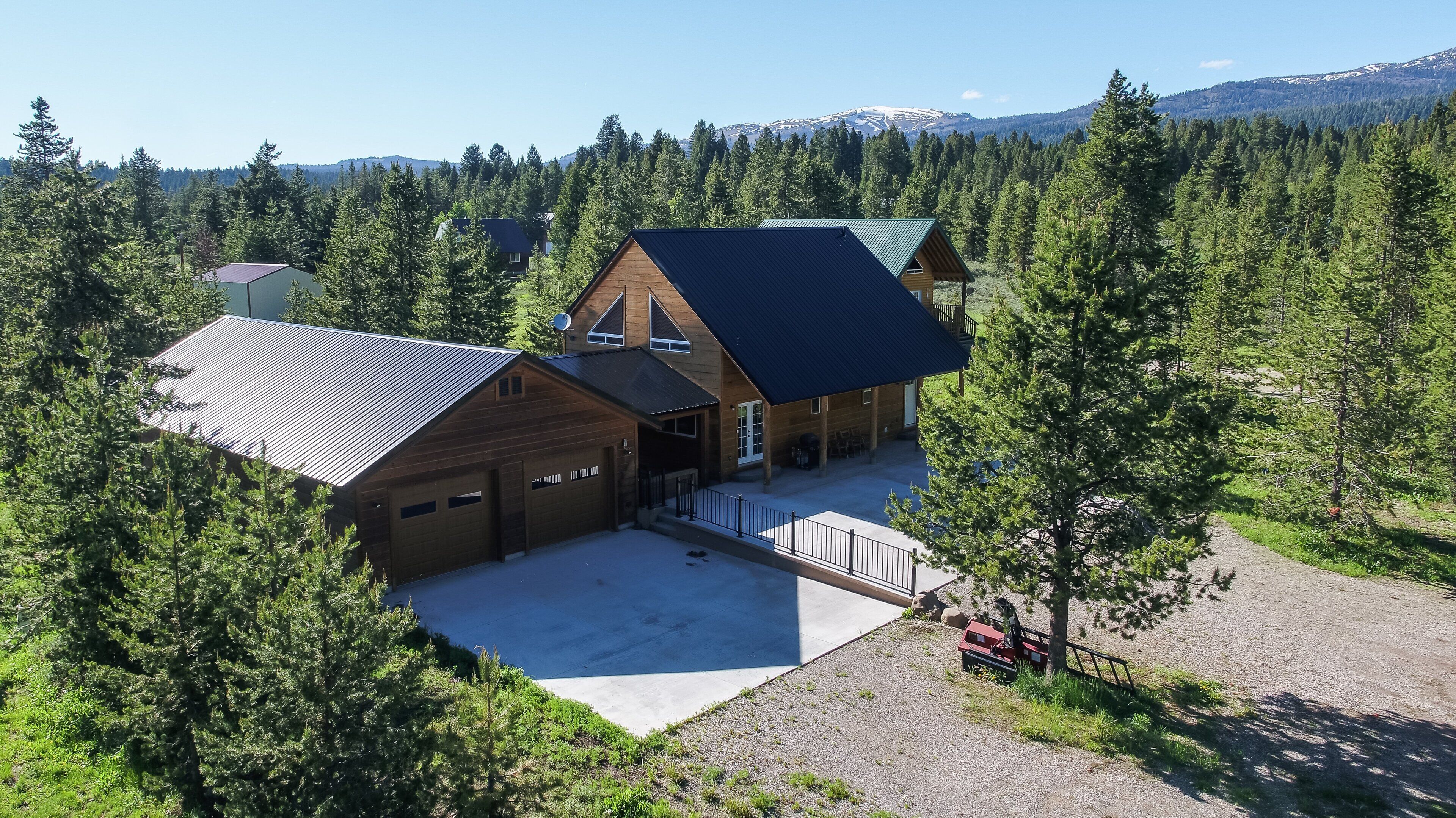 Une grande cabane en rondins avec un garage, entourée d’une forêt dense et avec des montagnes en arrière-plan.