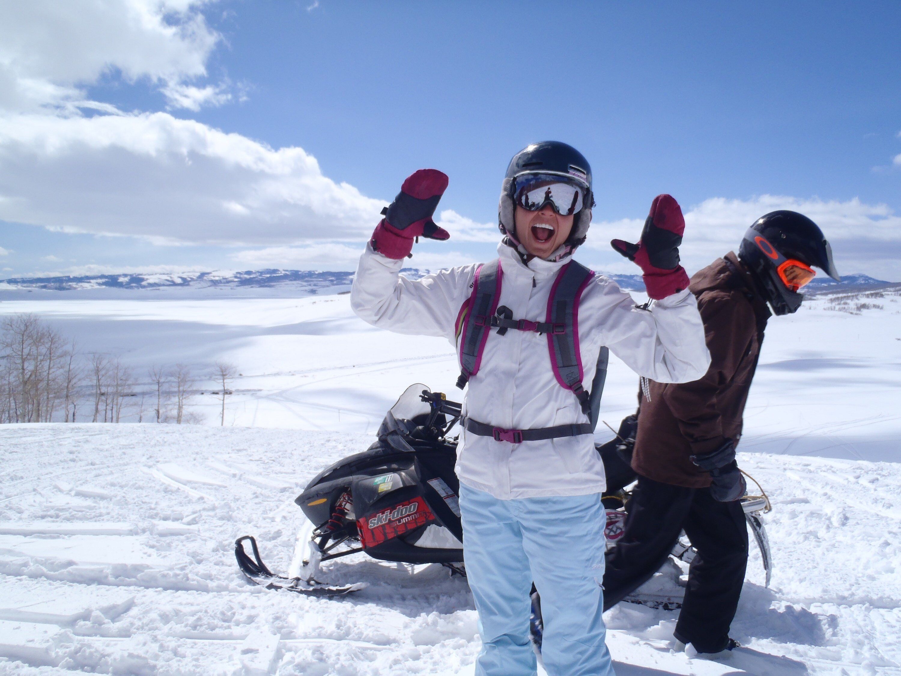 A snowy landscape with a snowmobile and two people wearing helmets and snow gear.
