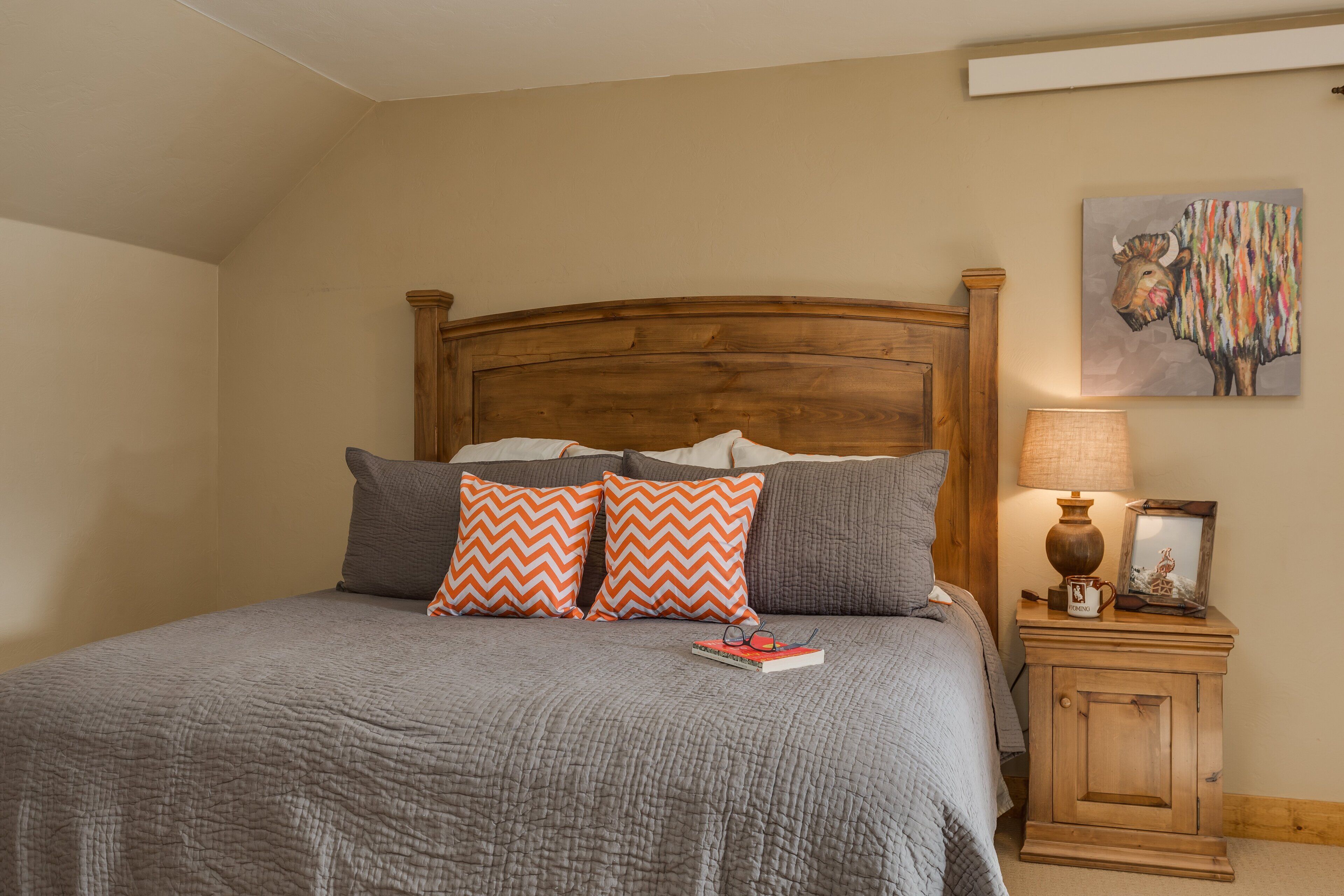 A bedroom with a wooden headboard, a gray quilted bedspread, two pillows with a zigzag pattern, a bedside table with a lamp, and a framed picture on the wall.