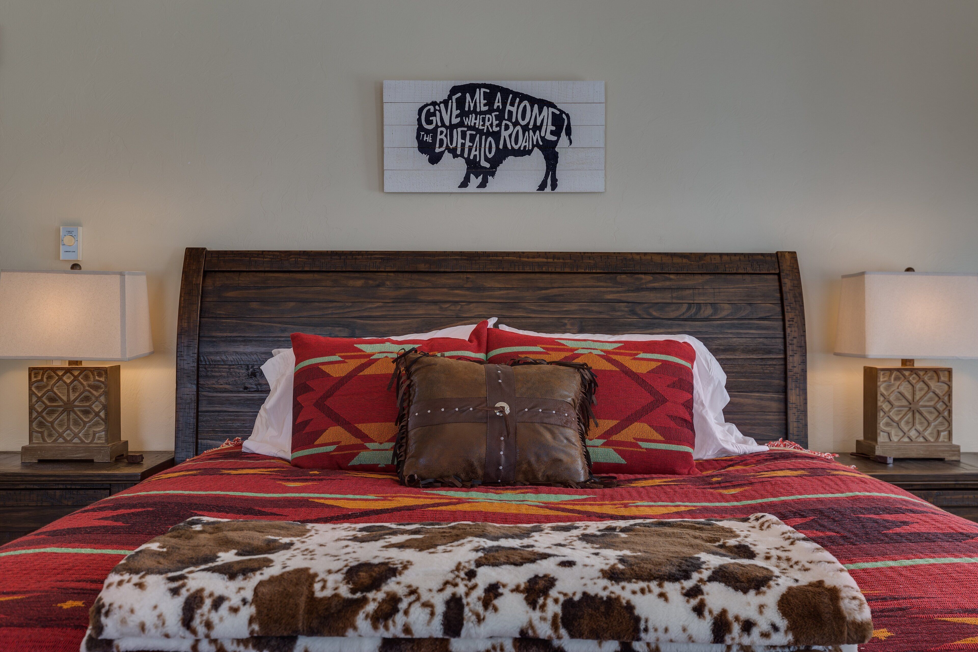A bedroom with a wooden headboard, a colorful bedspread, a cowhide throw, and a wall-mounted sign that reads "Give me a home where I roam."