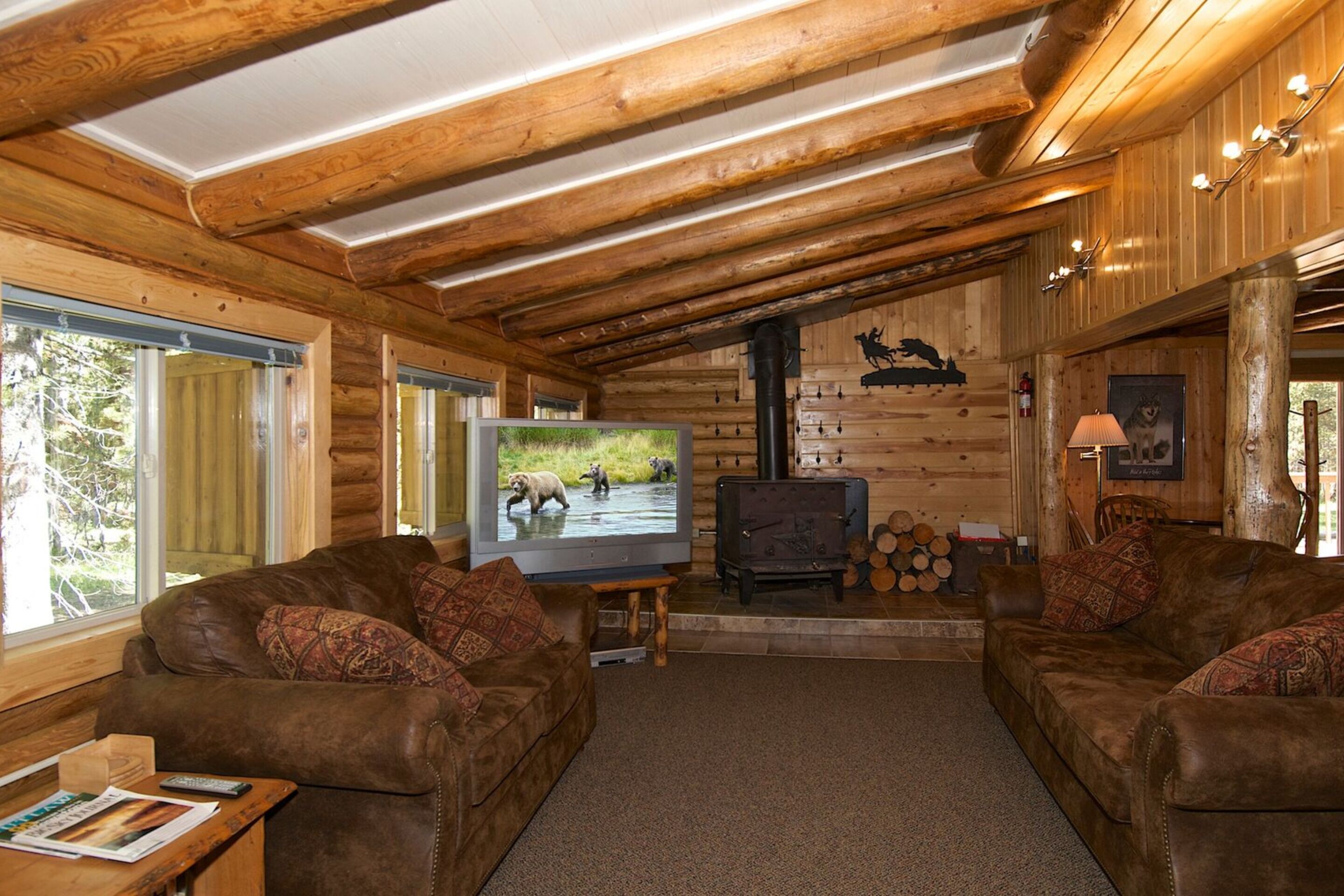 A log cabin interior with two leather sofas, a television, a fireplace, and a wooden wall with a bear silhouette.