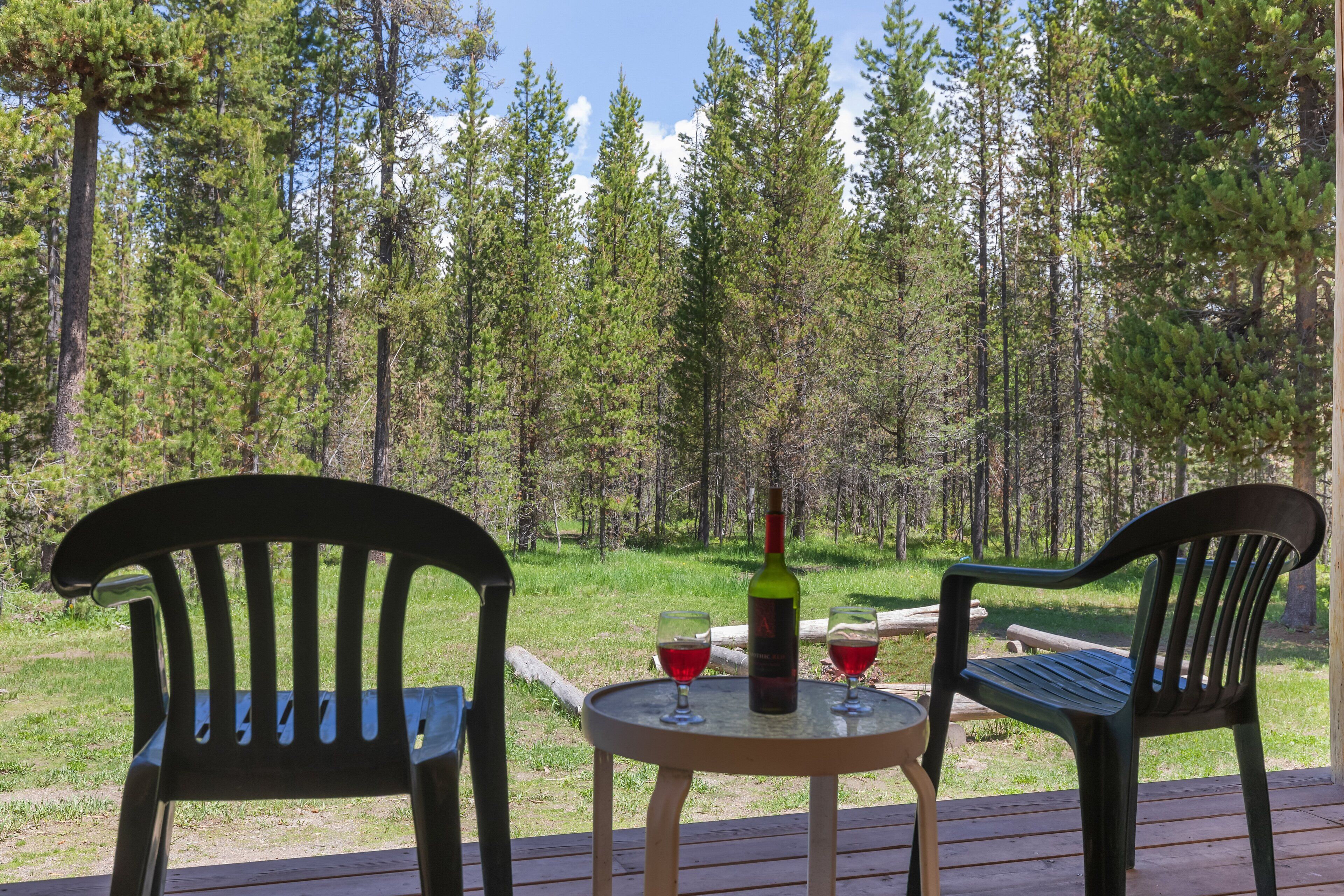 Zwei Stühle im Freien auf einer Holzterrasse mit einer Weinflasche und zwei Gläsern auf einem kleinen runden Tisch, mit Blick auf ein bewaldetes Gebiet.