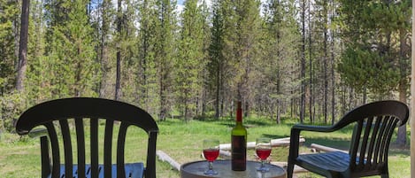 Zwei Stühle im Freien auf einer Holzterrasse mit einer Weinflasche und zwei Gläsern auf einem kleinen runden Tisch, mit Blick auf ein bewaldetes Gebiet.