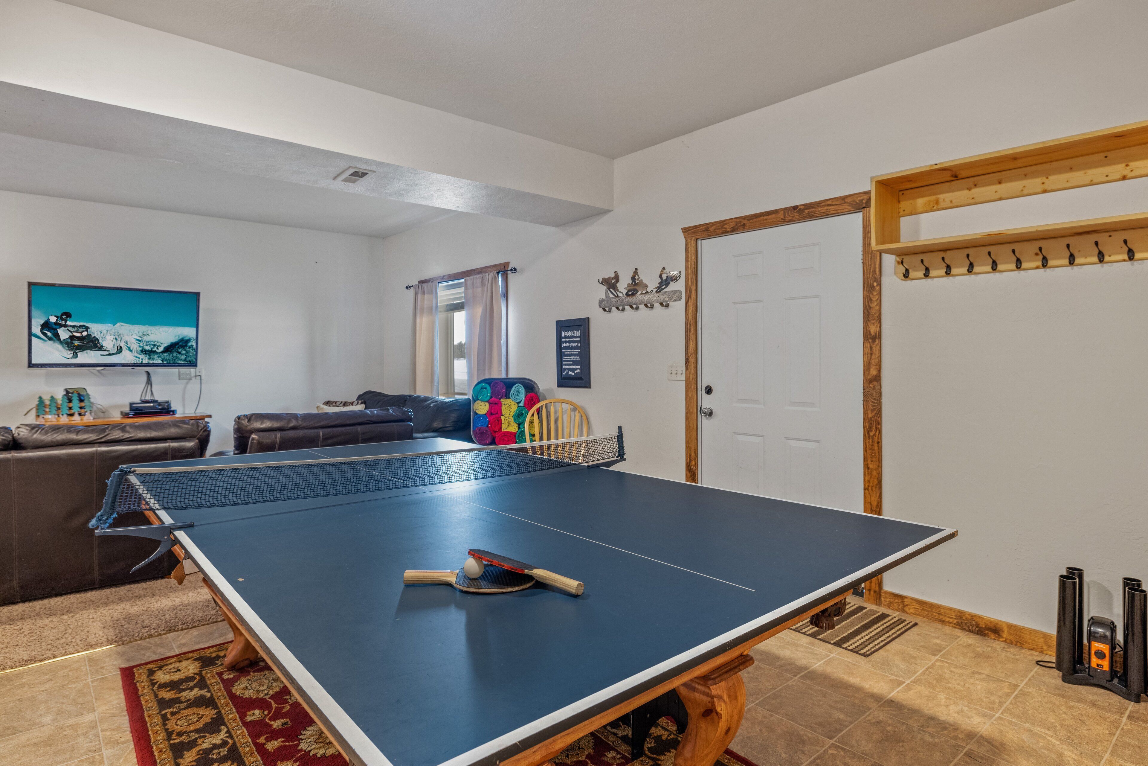 Une table de ping-pong dans un salon, avec un téléviseur, un canapé et une porte sur laquelle est accroché un porte-manteau.
