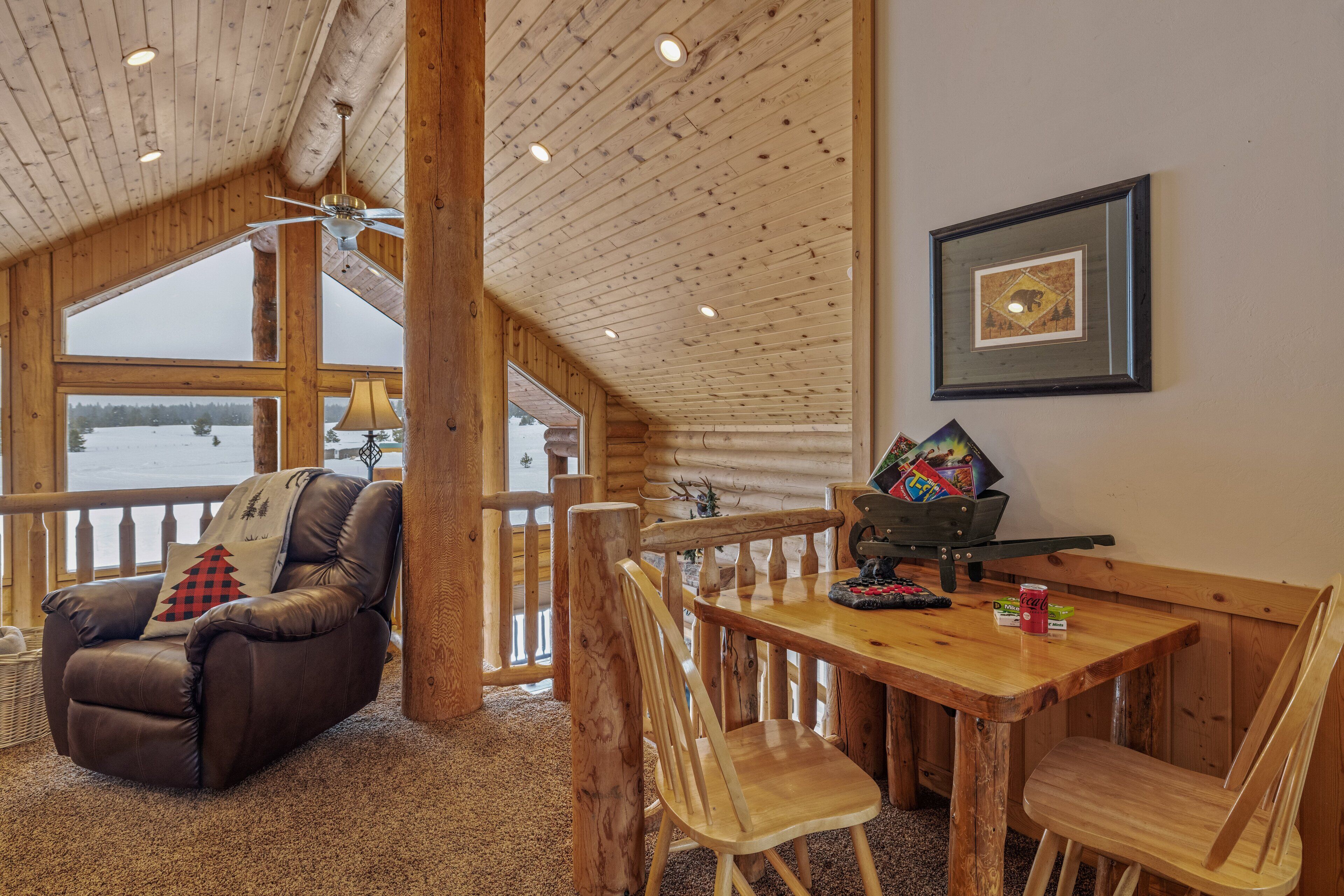 A cozy cabin interior with a wooden dining table, leather recliner, and a view of a snowy landscape through large windows.