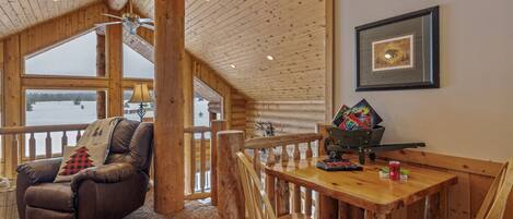 A cozy cabin interior with a wooden dining table, leather recliner, and a view of a snowy landscape through large windows.