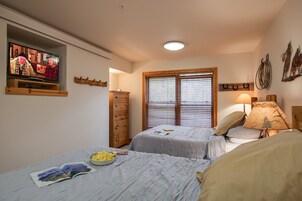 Un dormitorio con dos camas, un televisor en la pared, una ventana con persianas, una lámpara y un aparador de madera.