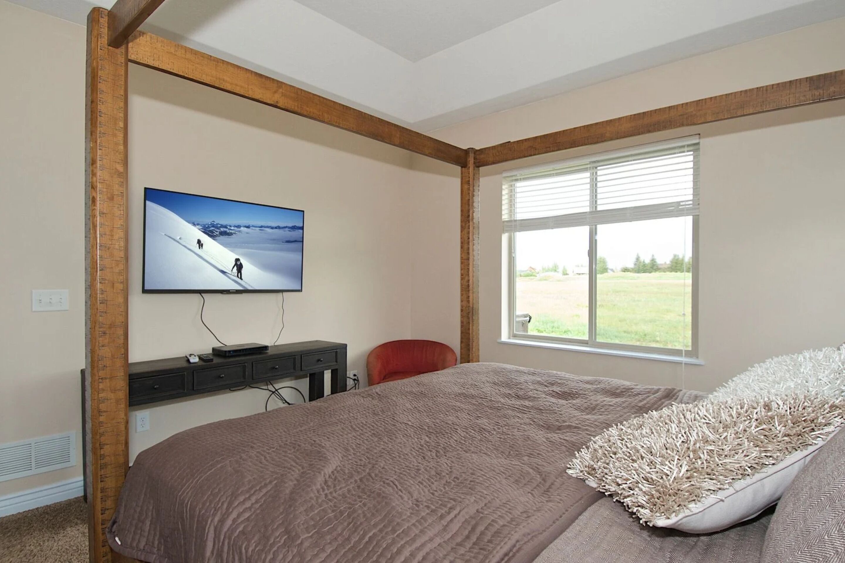 A bedroom with a large bed, a TV mounted on the wall, a window with blinds, and a desk with a chair.