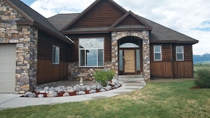 A two-story house with stone and wood siding, a stone chimney, and a front porch with a small garden. - Teton Harmony (Driggs)