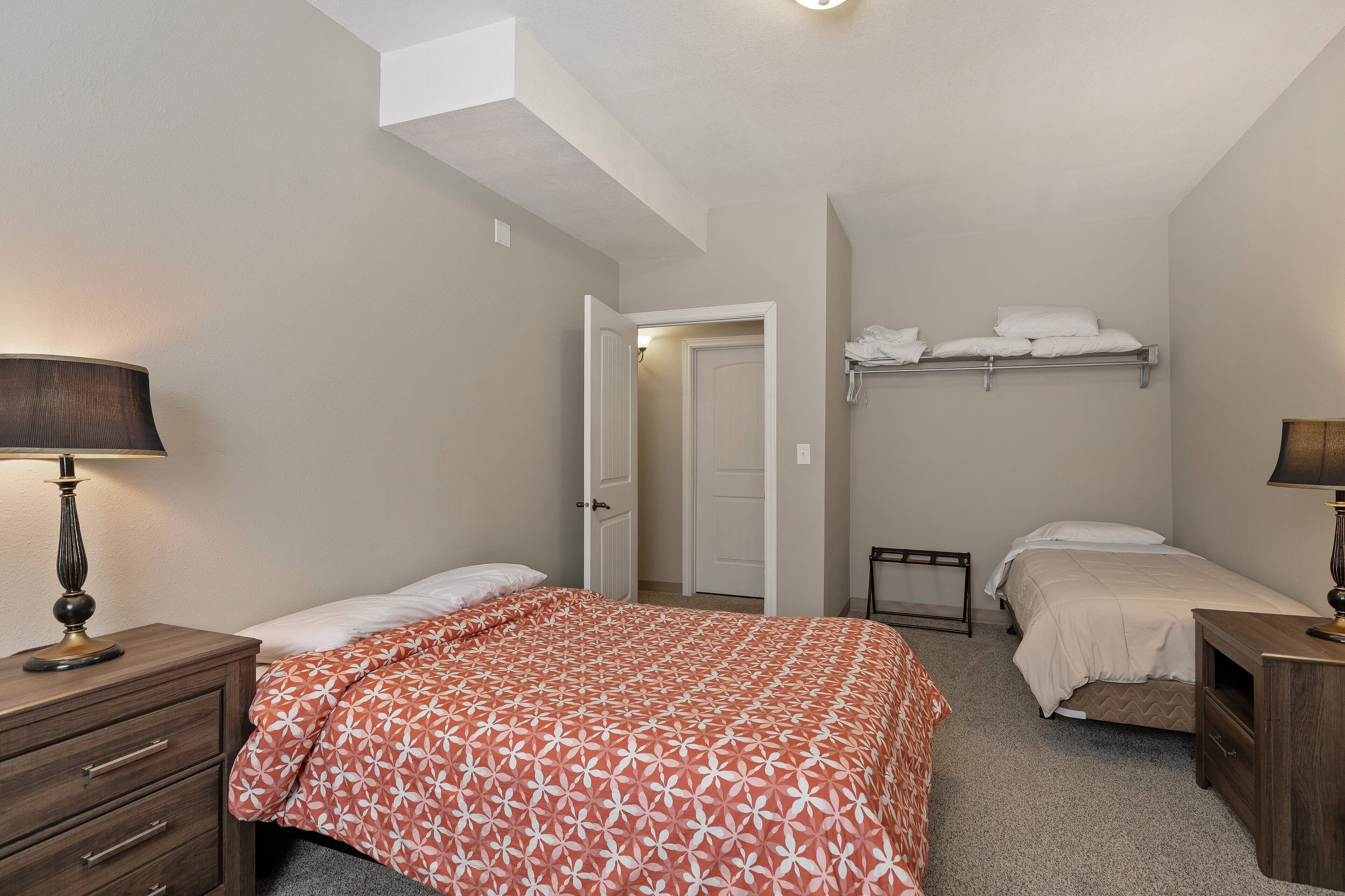 A bedroom with two beds, a nightstand with a lamp, a dresser, and a door leading to another room.