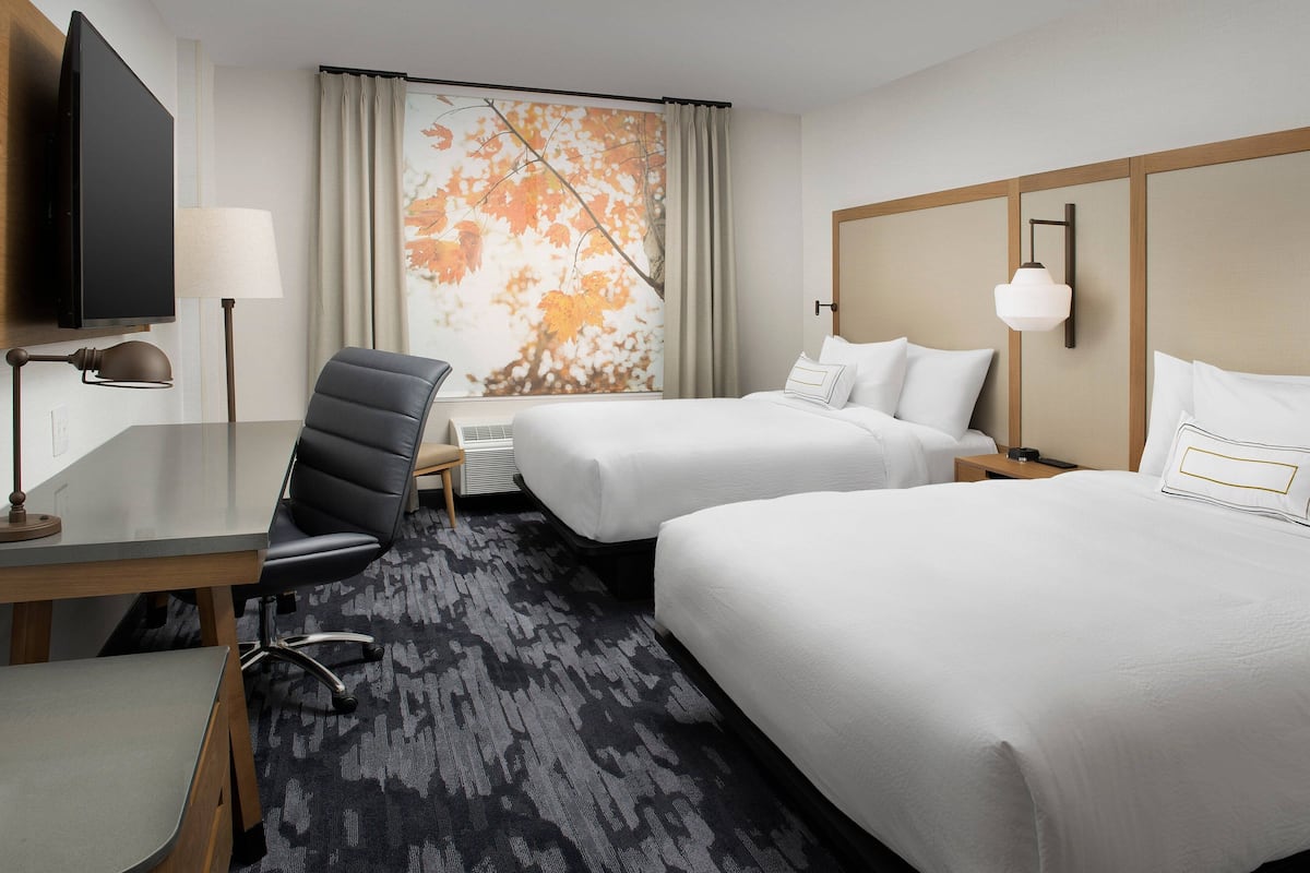 A hotel room with two beds, a desk with a chair, a television, and a large window with curtains.