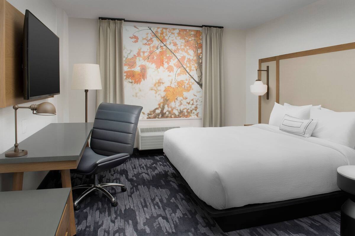 A hotel room with a bed, desk, chair, television, lamp, and a window with autumn leaves.