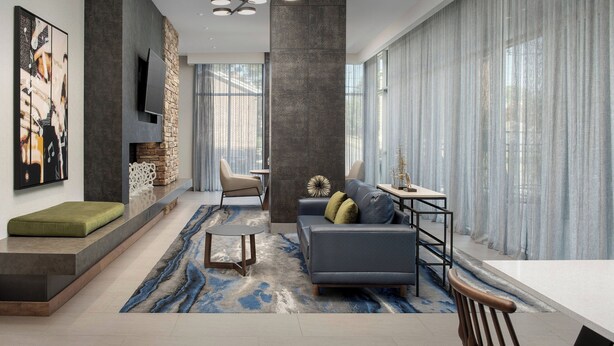 Una sala de estar moderna con un sofá gris, una mesa de centro y un televisor montado en una pared de piedra. Hay un ventanal con cortinas translúcidas y un tapete azul estampado en el suelo.